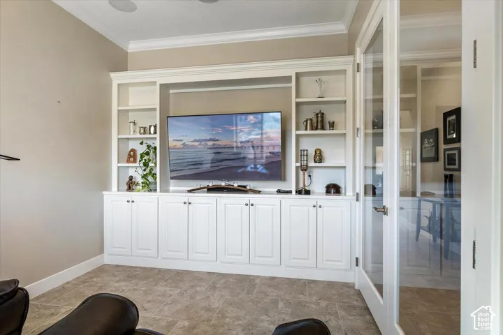 Custom built-in cabinetry featuring white shaker-style doors, integrated shelving, crown molding, and glass-paned French doors with brushed nickel hardware, complemented by neutral tile flooring