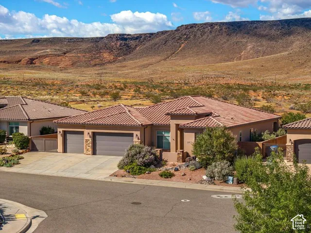 Single-level stucco exterior featuring a tiled roof, multi-car garage, stone accent pillars, and desert landscaping set against a mountain backdrop