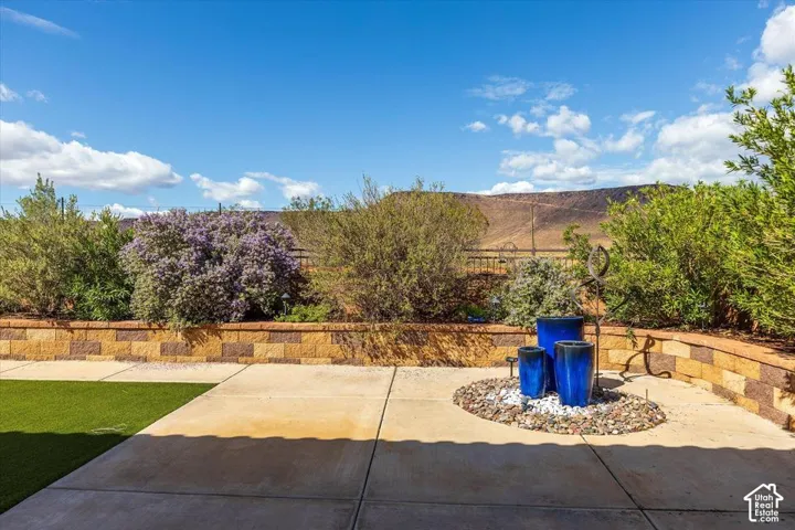 Backyard featuring concrete hardscaping, tiered block retaining wall, synthetic turf, and decorative rock garden with built-in water feature, set against a mountainous backdrop
