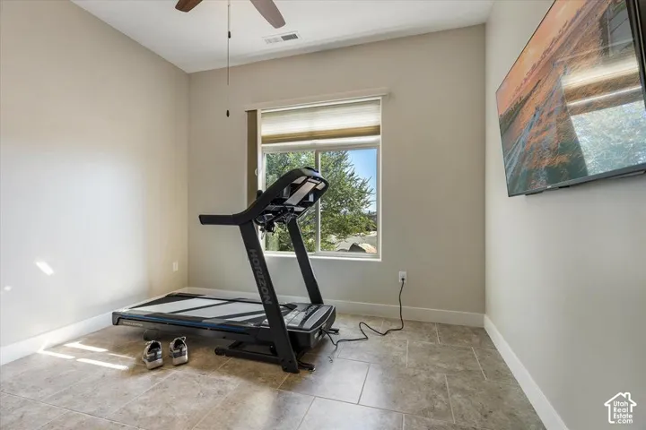 Neutral-toned room featuring stone-finish tile flooring, a large window with fitted pleated shades, a ceiling fan with integrated lighting, and a wall-mounted television bracket