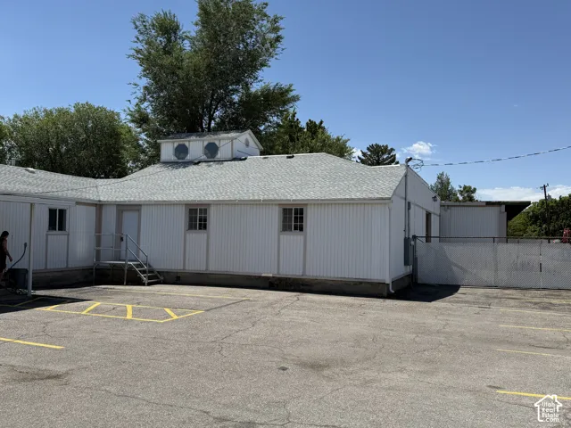 Exterior featuring white metal siding, shingled roof, decorative porthole windows, paved asphalt parking area, and metal perimeter fencing