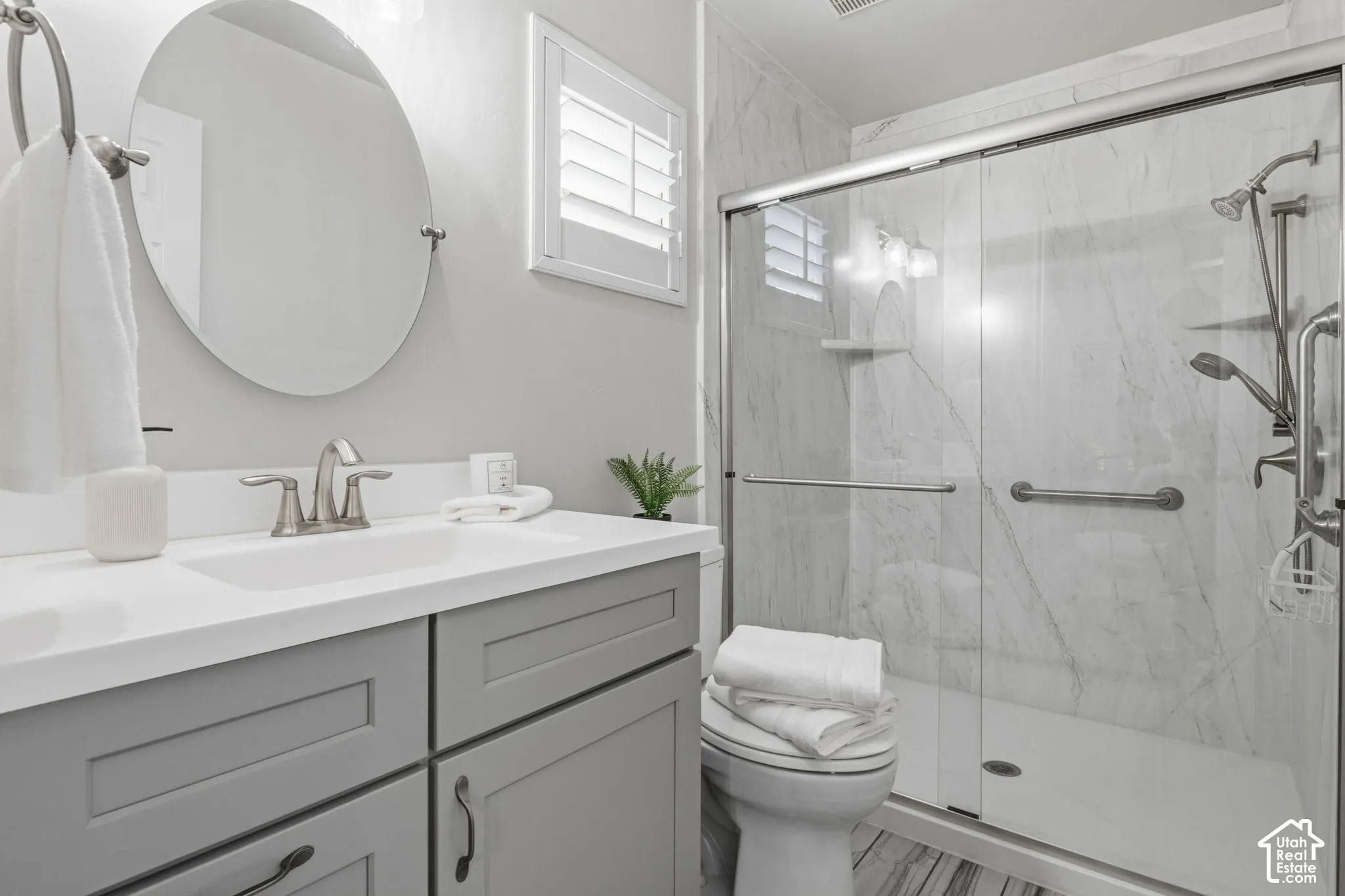 Grey shaker-style cabinetry with a white countertop, oval wall-mounted mirror, walk-in shower featuring a glass enclosure, large-format stone-patterned wall tile, and multi-setting shower fixtures