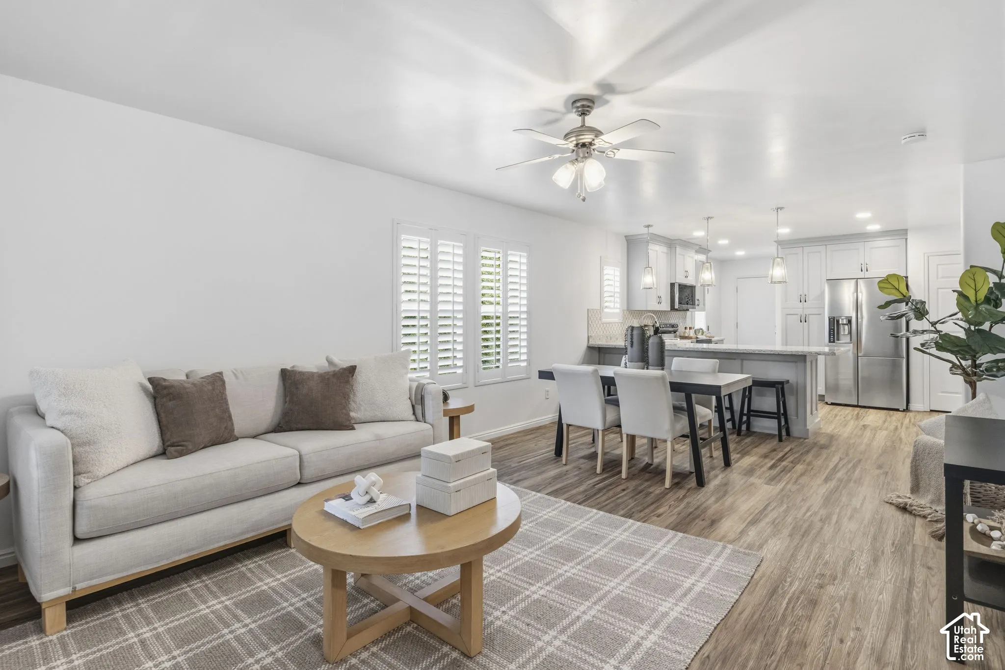 Open-concept interior featuring wood-finish flooring, plantation shutters, a ceiling fan with integrated lighting, recessed lighting, and kitchen cabinetry with a breakfast bar