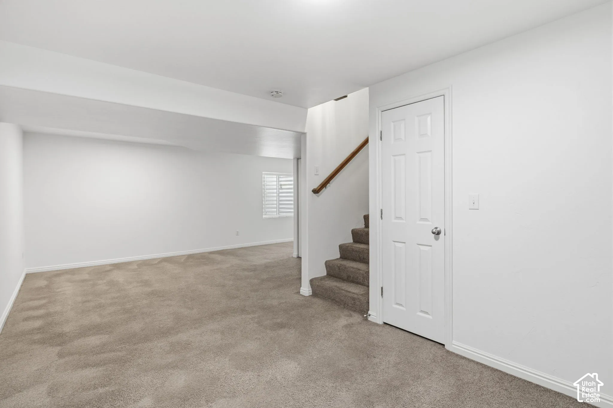 Open-concept interior featuring neutral wall-to-wall carpeting, white walls, shuttered window, and a paneled interior door with a wooden handrail
