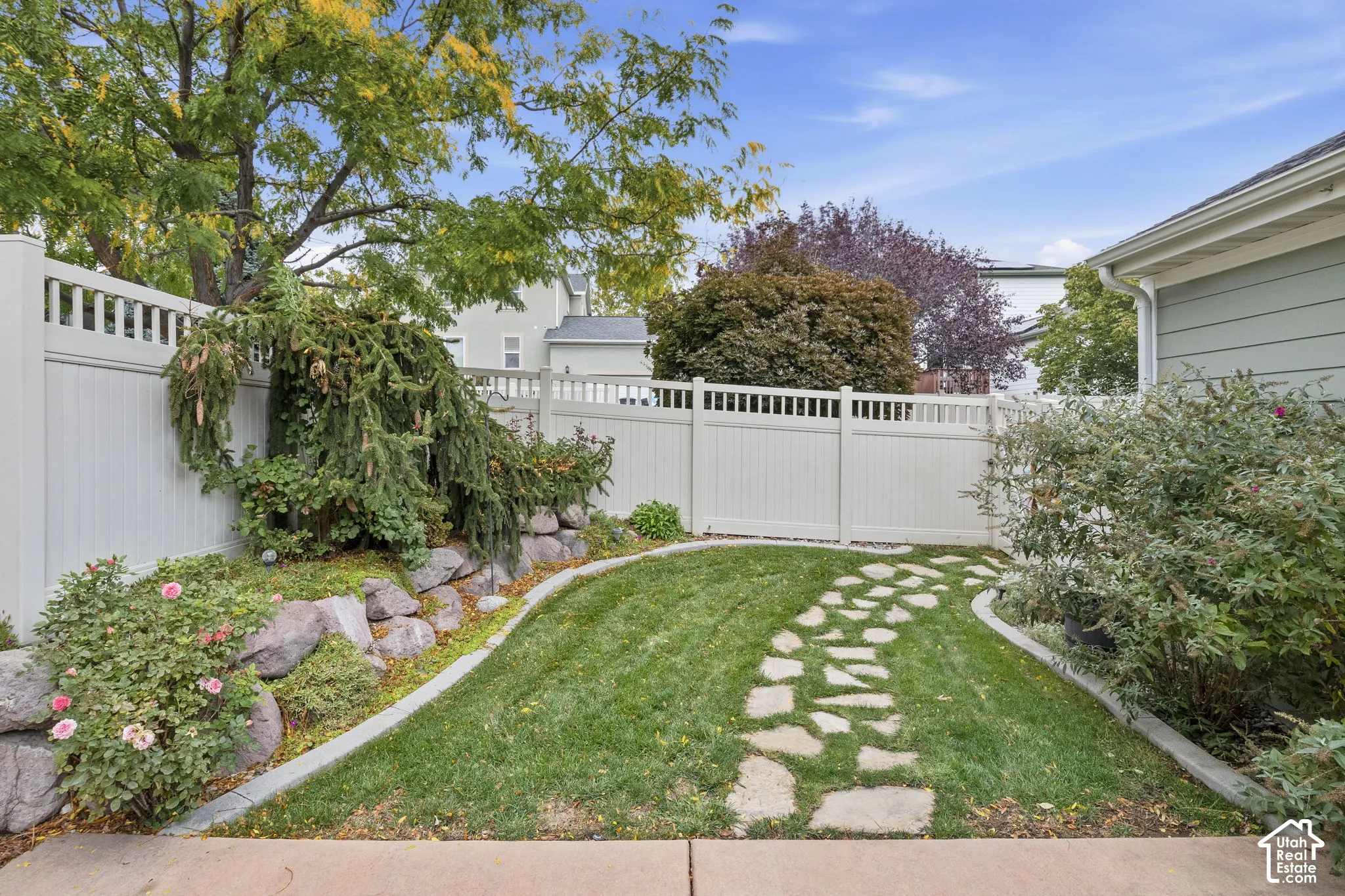 Backyard featuring a stone-paver walkway, manicured lawn, tiered natural rock landscaping, and a tall vinyl privacy fence