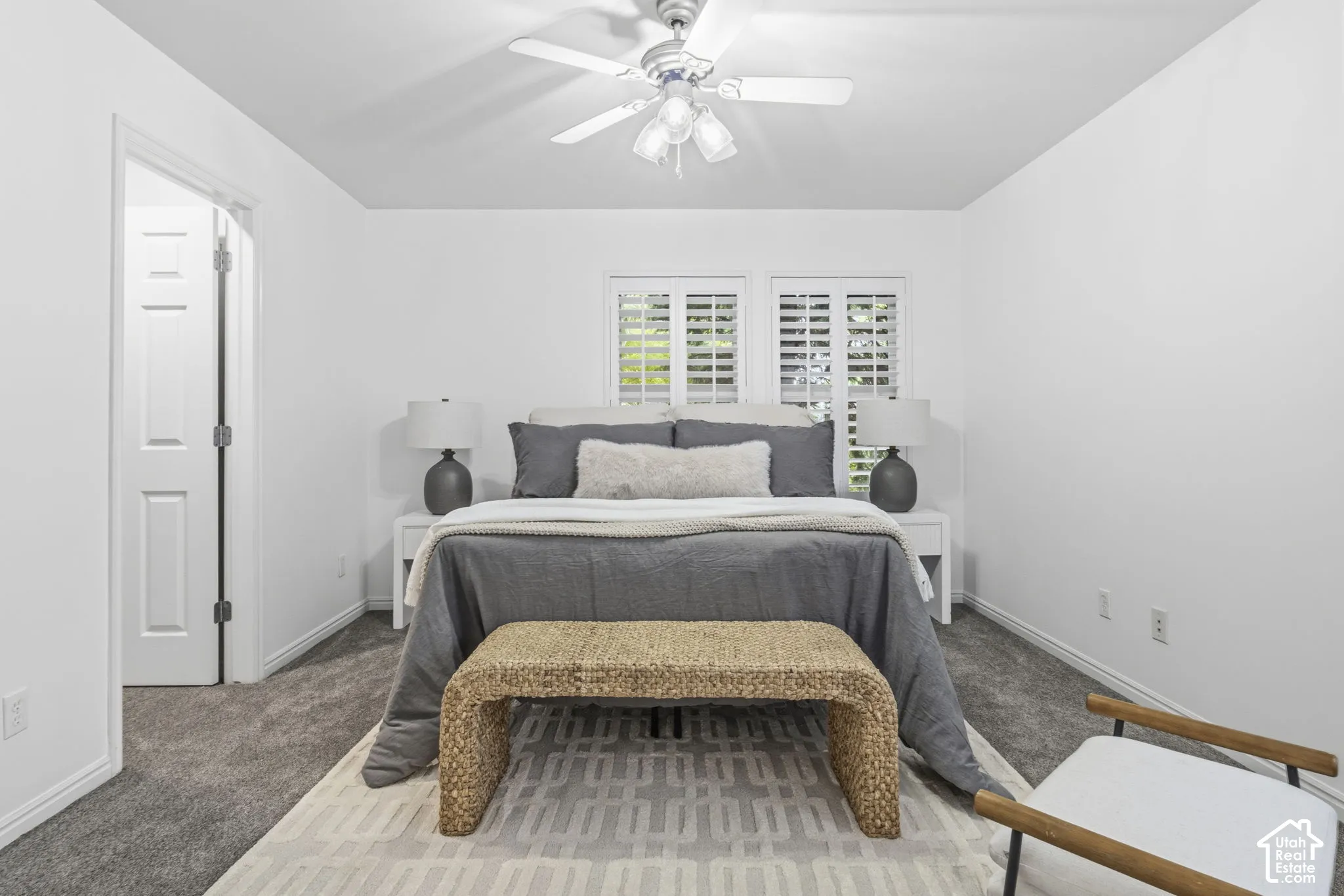 Neutral-toned interior featuring wall-to-wall carpeting, dual windows with interior plantation shutters, and a ceiling fan with integrated multi-bulb lighting