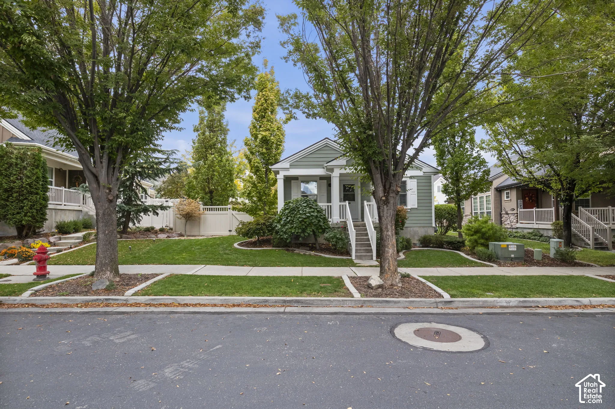 Single-story exterior featuring light-toned horizontal siding, a covered front porch, white column supports, and a gabled roofline, set within a landscape of mature deciduous trees and manicured lawn