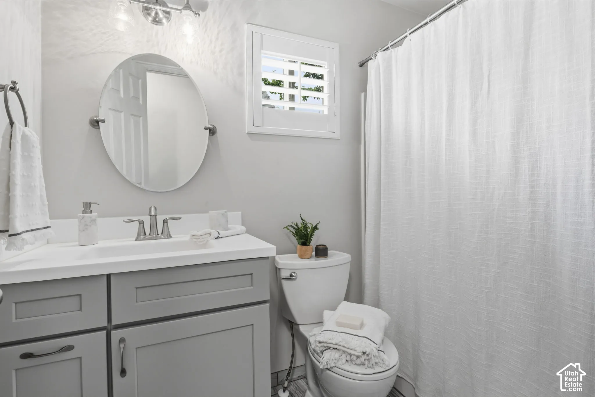 Bathroom featuring a gray shaker-style vanity with a solid surface countertop, an oval wall-mounted mirror, a two-handle metal faucet, and a window with plantation shutters