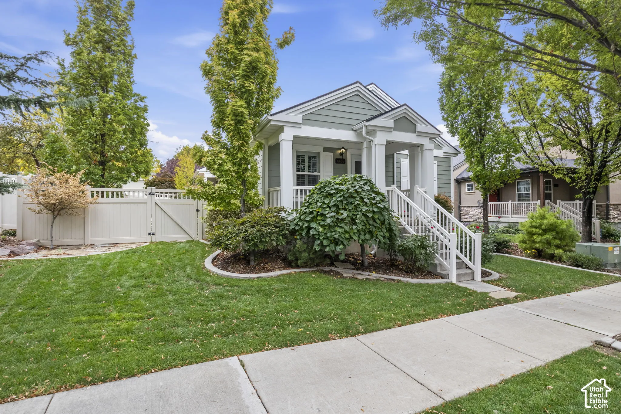 Craftsman-style exterior featuring sage green horizontal siding, a covered front porch with white railing, and a gabled roofline, complemented by landscaped garden beds and white vinyl privacy fencing