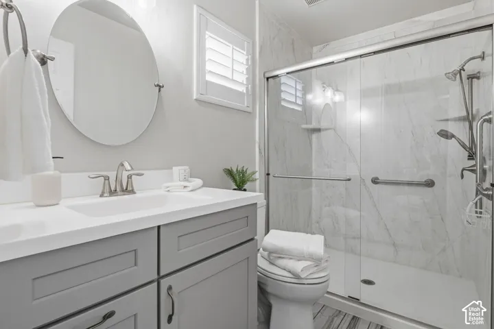 Grey shaker-style cabinetry with a white countertop, oval wall-mounted mirror, walk-in shower featuring a glass enclosure, large-format stone-patterned wall tile, and multi-setting shower fixtures