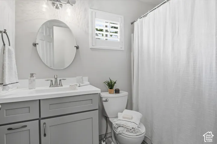 Bathroom featuring a gray shaker-style vanity with a solid surface countertop, an oval wall-mounted mirror, a two-handle metal faucet, and a window with plantation shutters