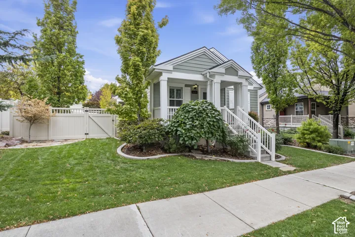 Craftsman-style exterior featuring sage green horizontal siding, a covered front porch with white railing, and a gabled roofline, complemented by landscaped garden beds and white vinyl privacy fencing