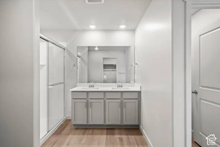 Dual-sink vanity with light gray cabinetry and integrated countertop, walk-in shower with glass enclosure, large vanity mirror, and wood-finish flooring