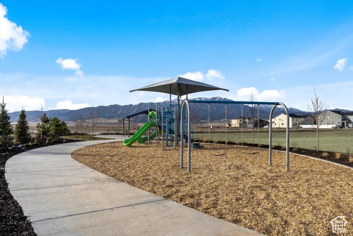 Community playground featuring structural play equipment, a covered canopy shade, mulch surfacing, and concrete pathways, set against a mountain backdrop