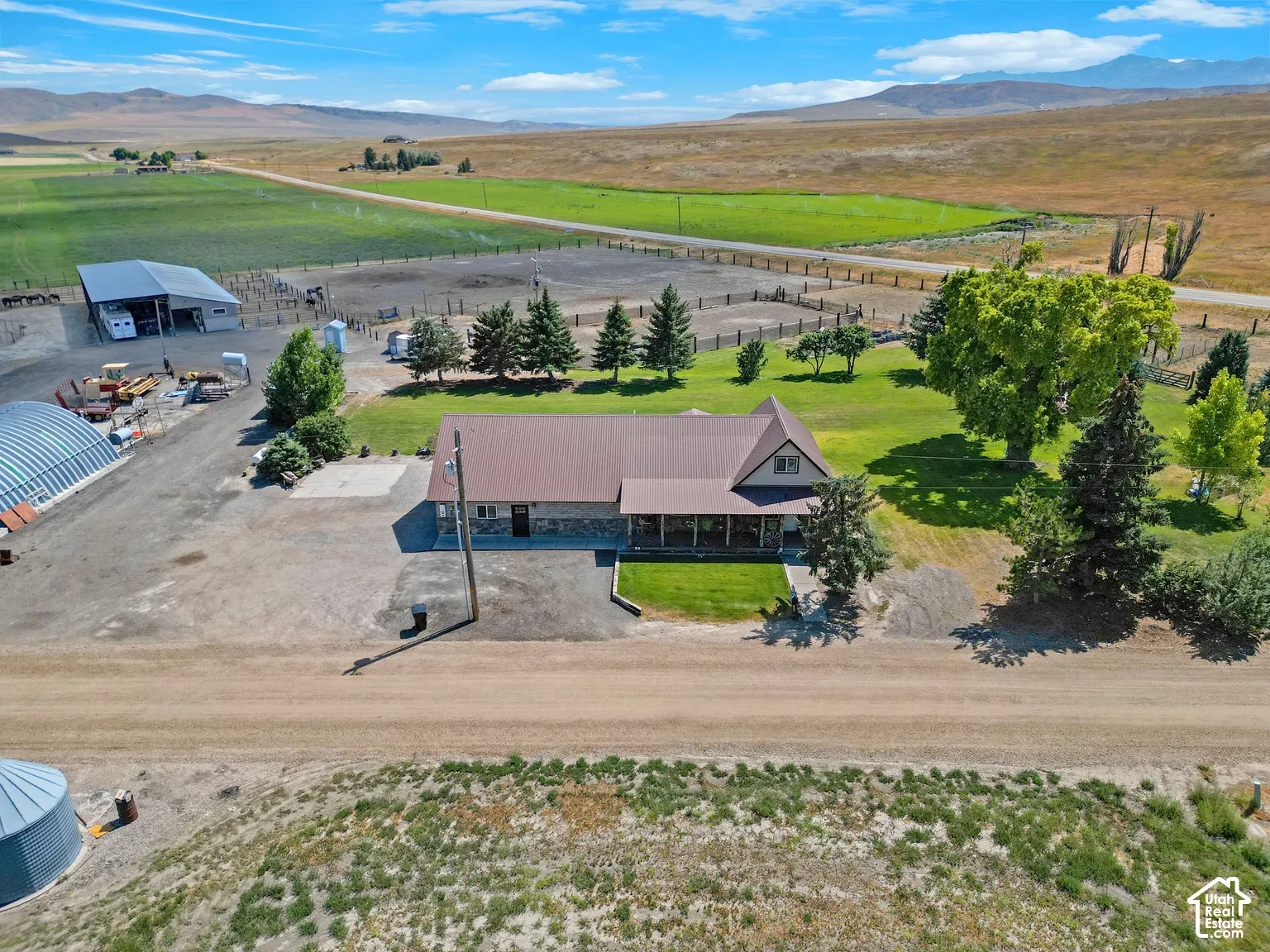Multi-structure property featuring a primary residence with stone siding, a metal roof, a covered front porch, and an auxiliary metal-sided outbuilding situated on expansive grounds with mature trees and mountain scenery