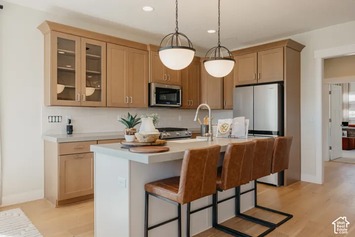 Kitchen featuring light-tone cabinetry with glass-front display uppers, expansive center island with seating overhang, white subway tile backsplash, stainless steel appliances, and wood-finish flooring