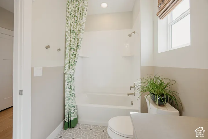 Full bathroom featuring a tub-and-shower combination with white surround, patterned tile flooring, recessed lighting, and a window with a woven shade