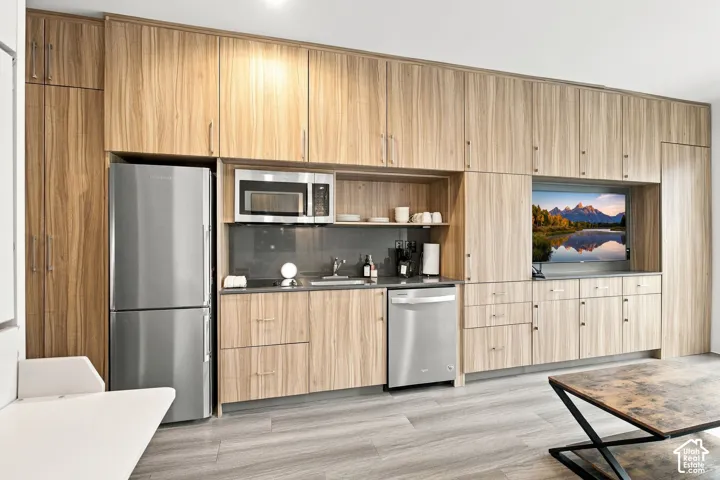 Full-height wood-finish cabinetry, integrated stainless steel appliances, dark grey solid-surface backsplash, wood-finish flooring, and built-in television alcove