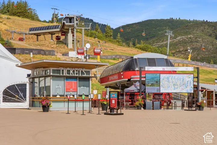 Paved plaza featuring a ticket kiosk, ski lift terminal, gondola station, and mountain terrain backdrop