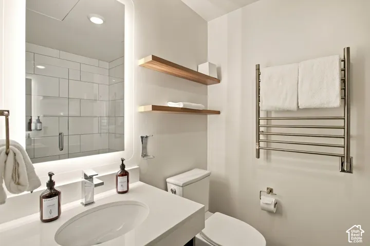 Bathroom featuring an integrated vanity with a quartz countertop, backlit mirror, wall-mounted heated towel rack, wood-finish floating shelving, and a glass-enclosed shower with rectangular tile