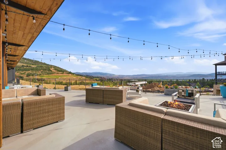 Expansive elevated terrace featuring a wood-clad overhang, integrated overhead string lighting, a stone fire pit, and metal railing with unobstructed mountain scenery