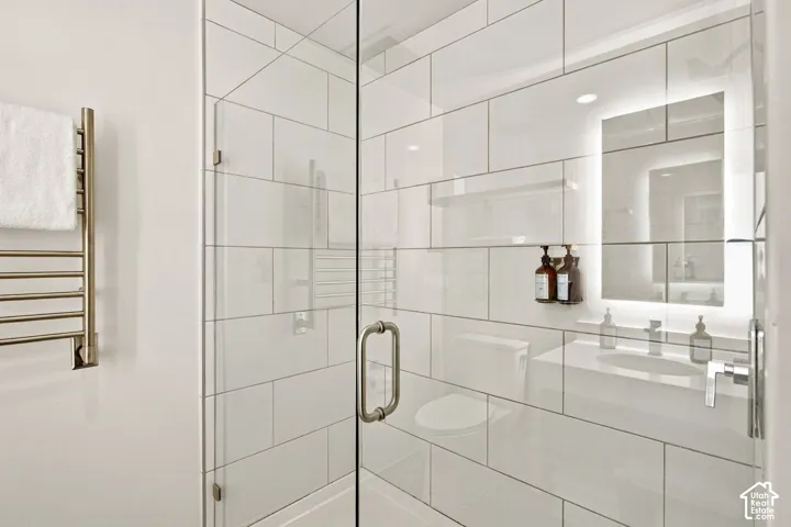Walk-in shower featuring white subway tile, frameless glass enclosure, wall-mounted heated towel rack, illuminated vanity mirror, and recessed lighting