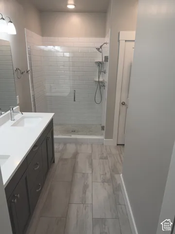 Double vanity featuring a white countertop with integrated sinks, dark cabinetry, walk-in shower with white subway tile surround, and wood-finish flooring