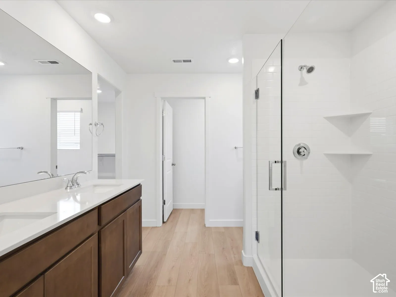 **DISCLAIMER** Interior package may vary. Bathroom featuring wood-finish cabinetry, white countertop with dual integrated sinks, full-width mirror, wood-finish flooring, and a walk-in shower with glass enclosure and white tile surround