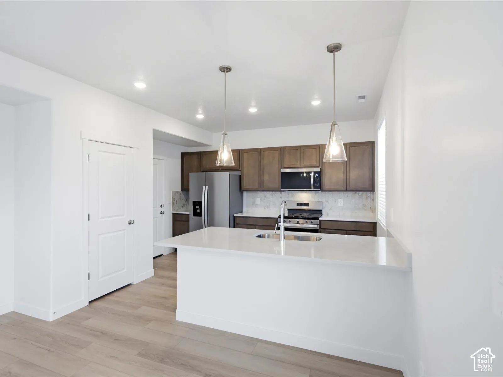 **DISCLAIMER** Interior package may vary. Kitchen featuring wood-finish cabinetry, quartz countertops, a central island with an integrated sink, stainless steel appliances, and pendant lighting