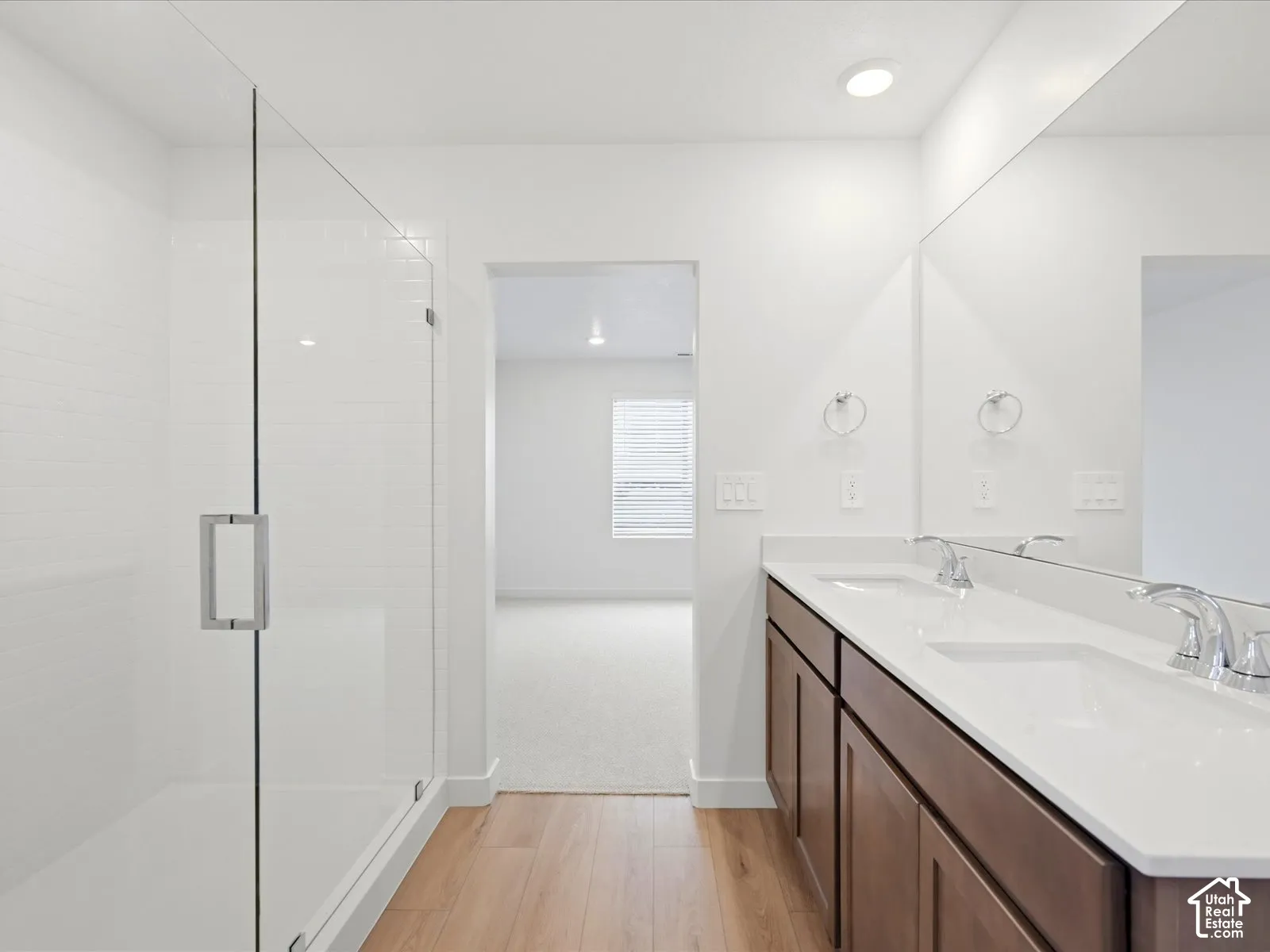 **DISCLAIMER** Interior package may vary. Double vanity with white solid-surface countertop, wood-finish cabinetry, full-width mirror, and glass-enclosed shower with subway tile surround, featuring wood-finish flooring