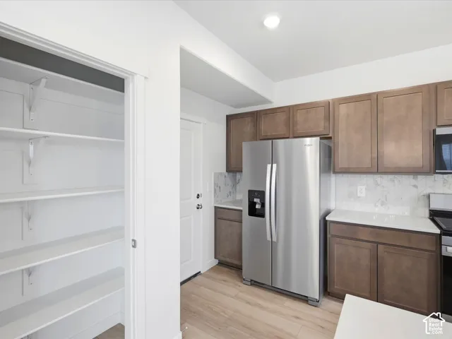 **DISCLAIMER** Interior package may vary. Kitchen featuring stained wood cabinetry, quartz countertops, white stone-finish backsplash, stainless steel refrigerator, and a walk-in pantry with built-in shelving