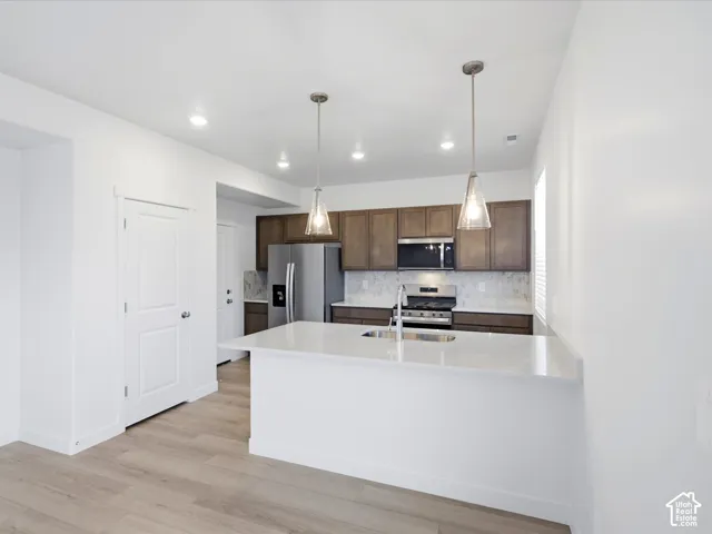 **DISCLAIMER** Interior package may vary. Kitchen featuring wood-finish cabinetry, quartz countertops, a central island with an integrated sink, stainless steel appliances, and pendant lighting