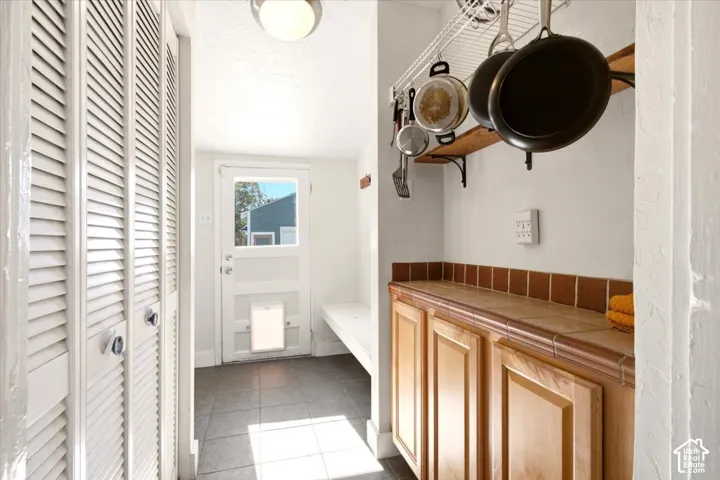 Utility area featuring tile flooring, built-in wooden cabinetry with tiled countertops, louvered closet doors, and a glass-paneled exterior door