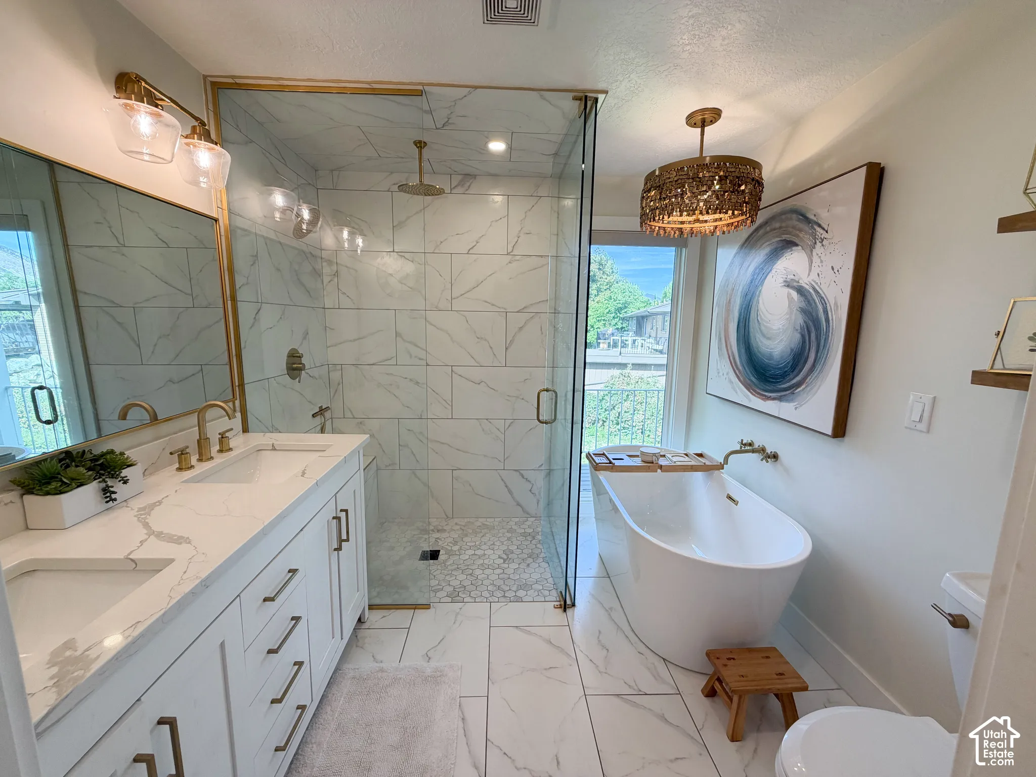 Dual-sink vanity with quartz countertops, glass-enclosed walk-in shower with floor-to-ceiling tile, freestanding soaking tub, and brass-tone fixtures