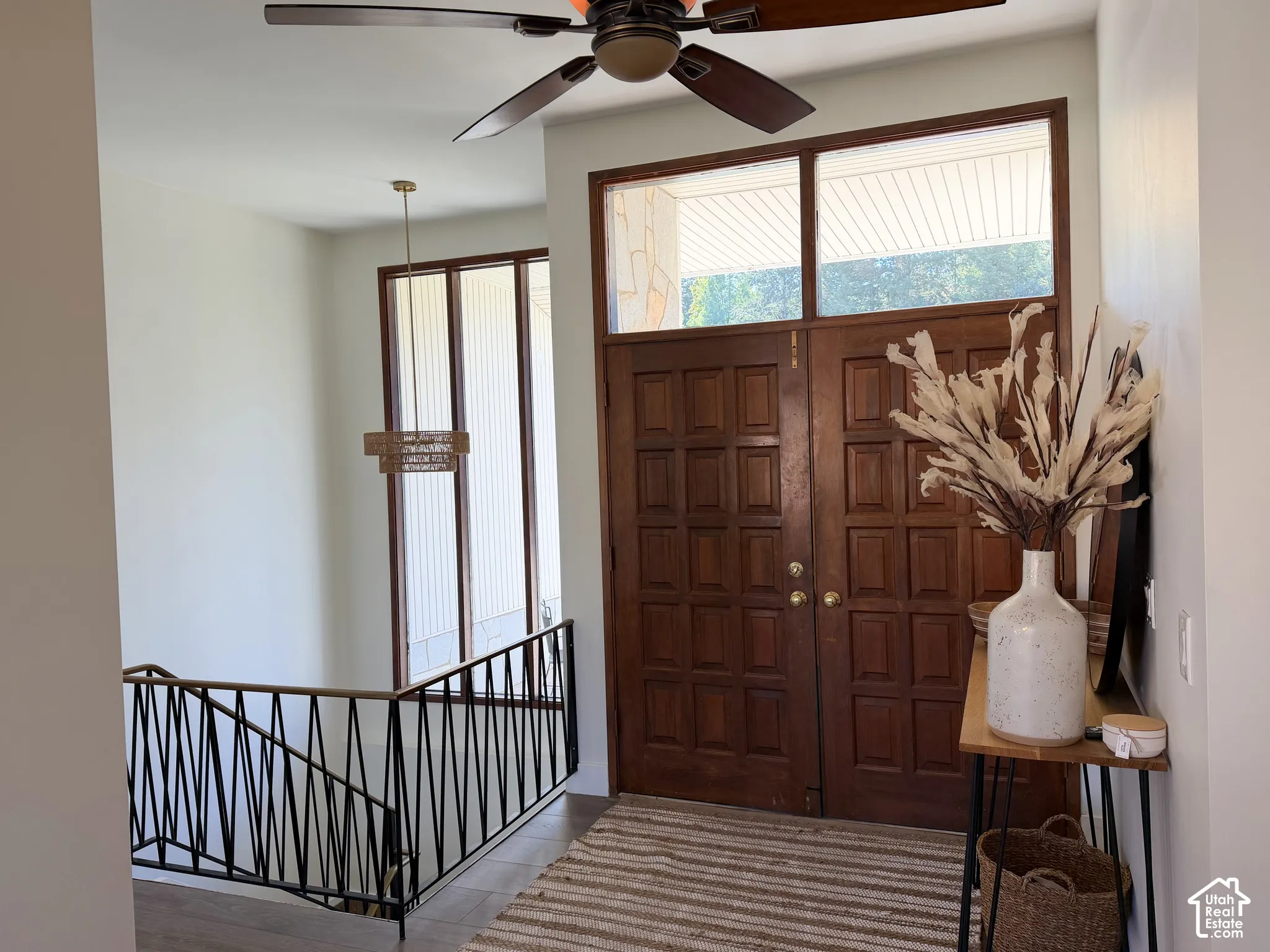 Double entry doors featuring raised panels and a transom window, geometric metal railing, large floor-to-ceiling windows, and a ceiling fan with integrated lighting