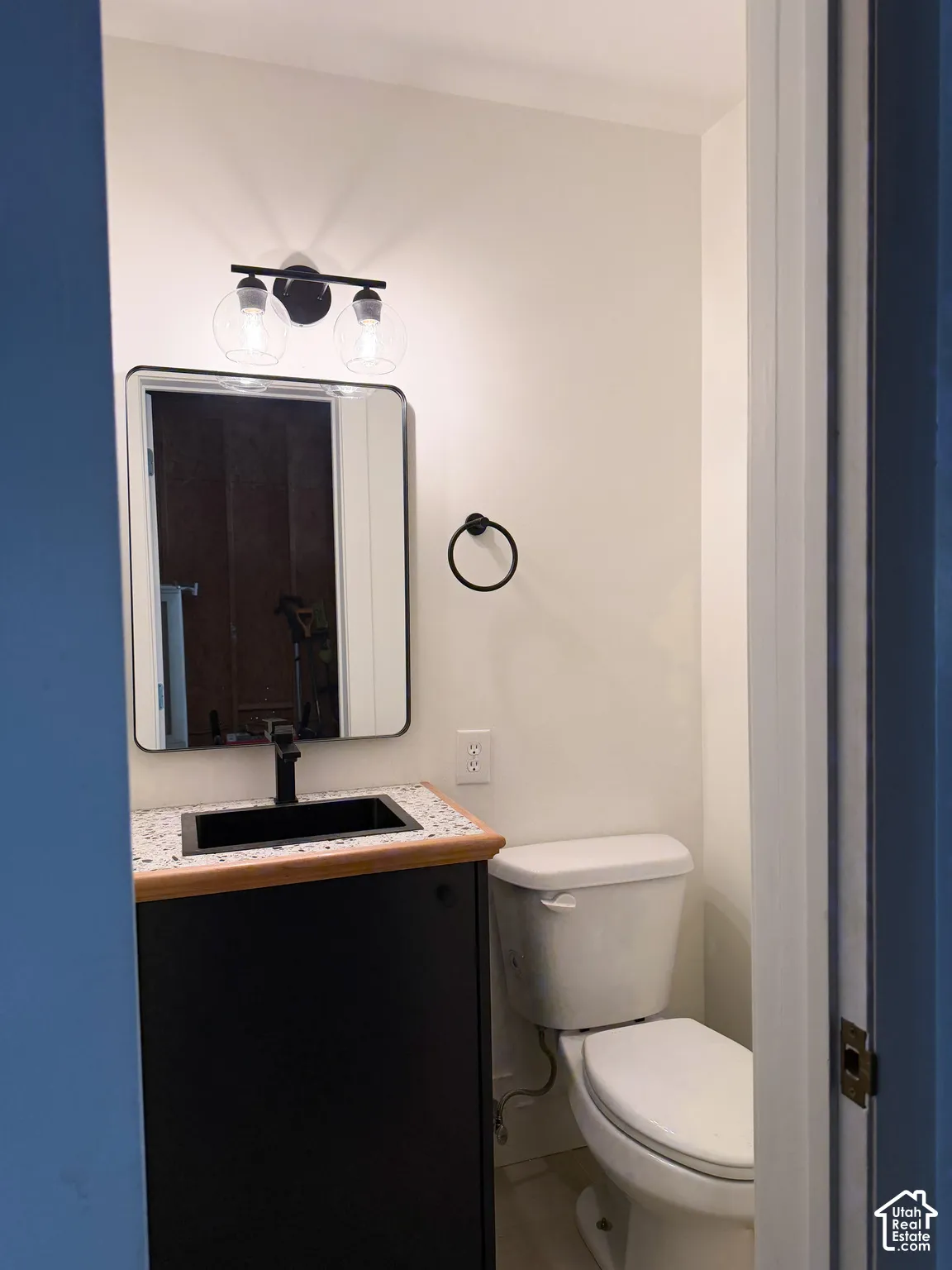 Garage bathroom - featuring a speckled countertop, integrated rectangular sink, matte black faucet, and modern wall-mounted light fixture, paired with a rounded-corner mirror and coordinating matte black towel ring