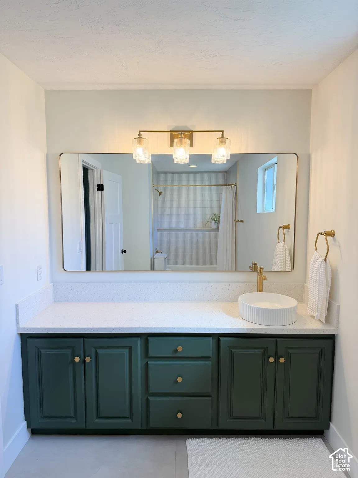 Deep green custom cabinetry paired with a white solid-surface countertop, a cylindrical fluted vessel sink, brass-tone hardware, and a three-light vanity fixture