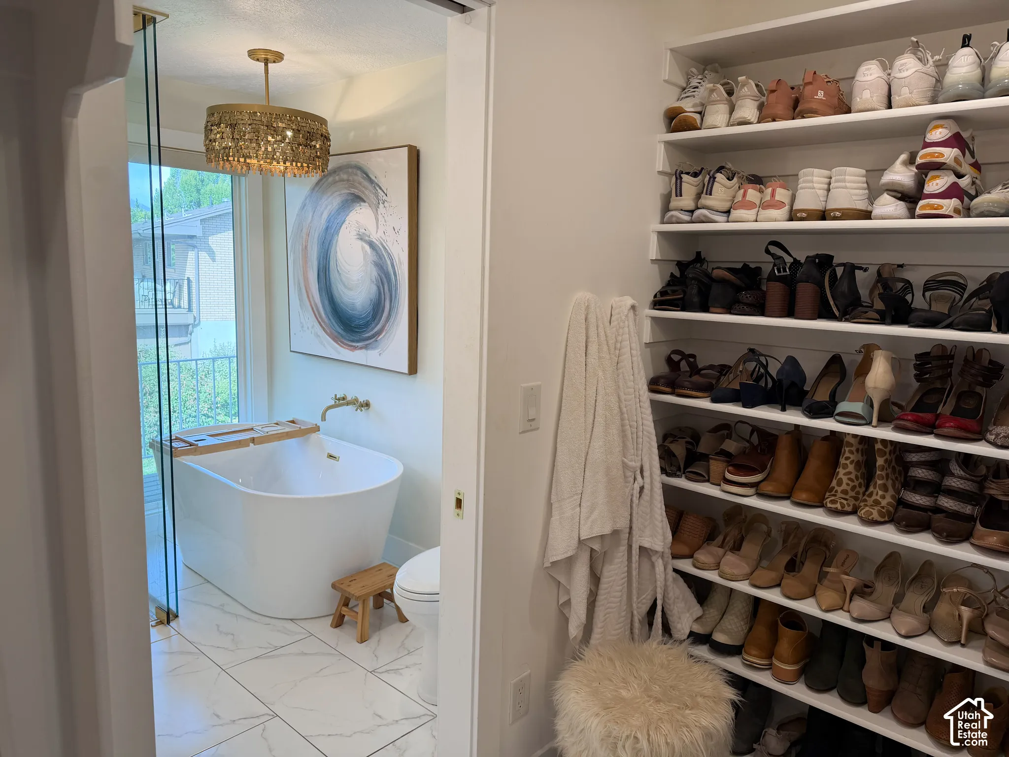 Freestanding soaking tub with wall-mounted brass faucet, floor-to-ceiling glass enclosure, tile flooring with stone-pattern finish, gold-toned drum chandelier, and built-in custom shelving
