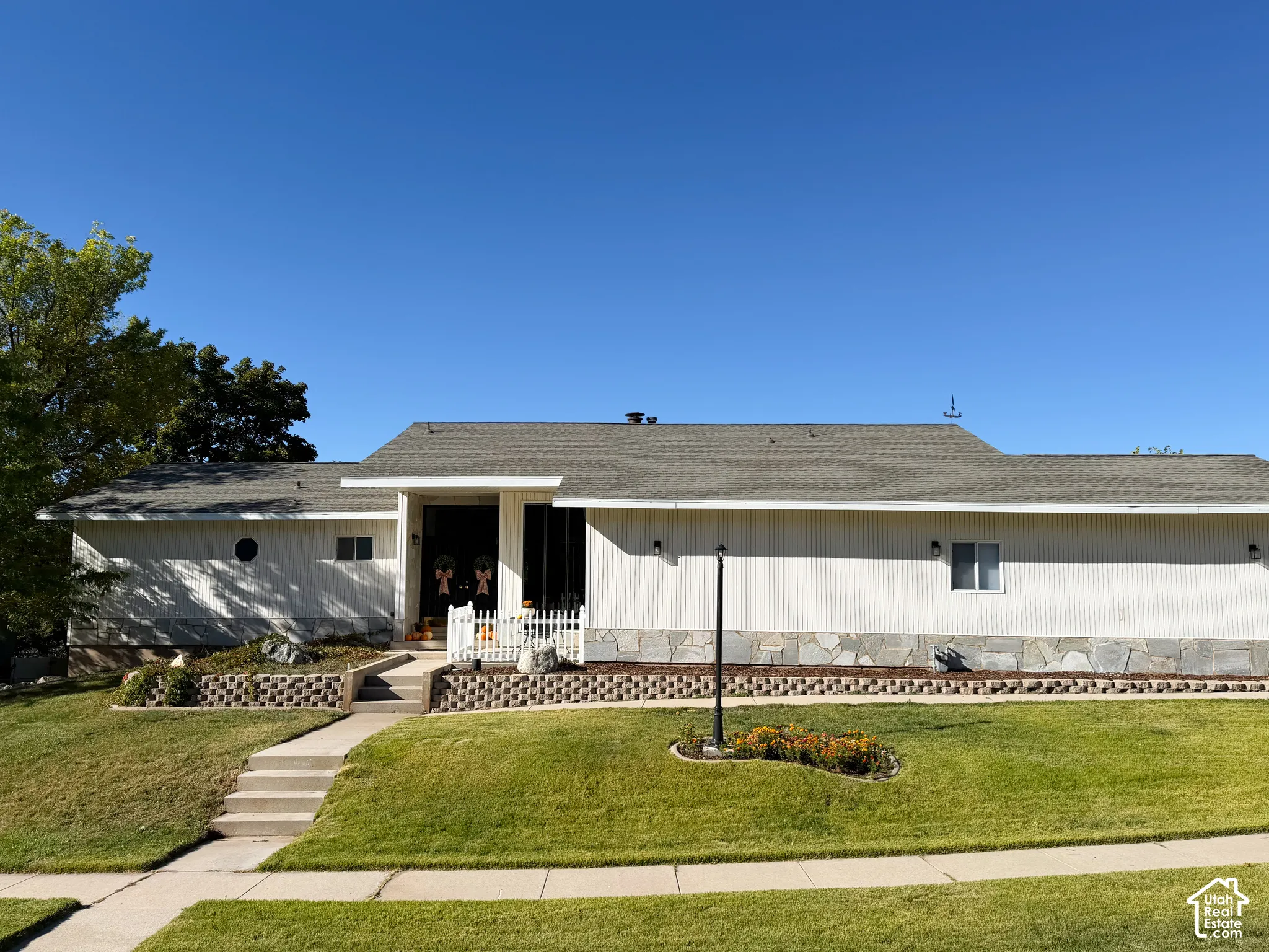 Single-story exterior featuring light-toned vertical siding, stone masonry skirting, an asphalt shingle roof, a concrete walkway, and a tiered retaining wall