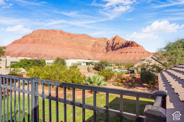 Elevated outdoor space with black metal railing, featuring a lawn, mature desert landscaping, stone retaining wall, and prominent red rock mountain backdrop