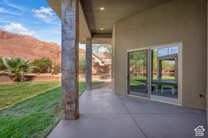 Covered concrete patio featuring stone-clad support columns, recessed lighting, and large sliding glass doors overlooking a landscape with a mountainous backdrop