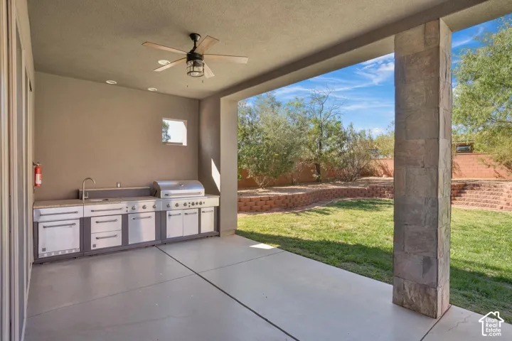 Covered patio featuring an outdoor kitchen with stainless steel appliances, stone-clad pillar, ceiling fan, and tiered retaining wall landscaping