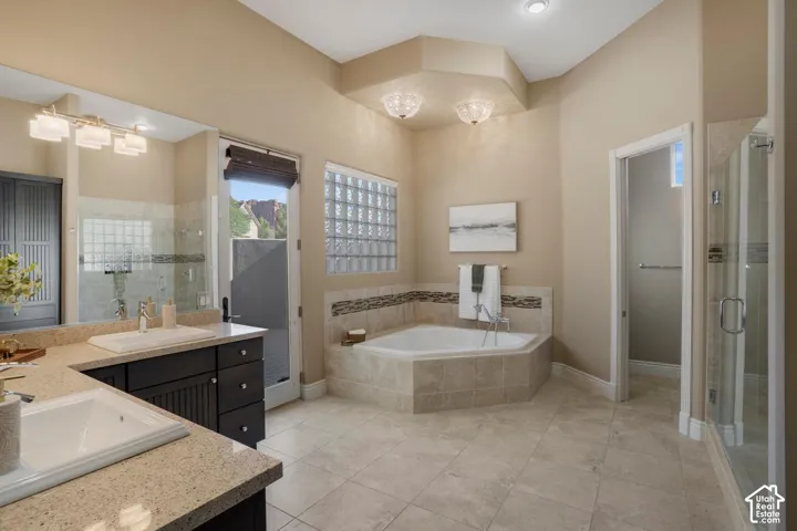 Dual-sink vanity with stone-finish countertops, corner soaking tub with tile surround, glass-block window, walk-in glass-enclosed shower, and tile flooring