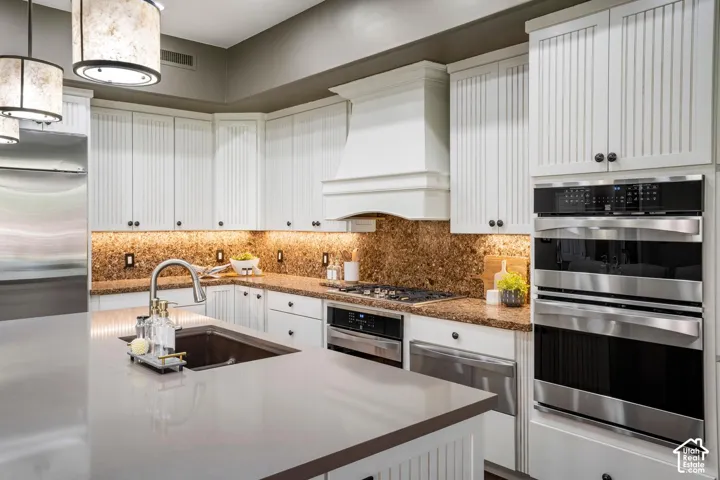 Kitchen featuring white vertical-paneled cabinetry, a large central island with a solid-surface countertop, integrated stainless steel double wall ovens, a custom range hood, and task lighting