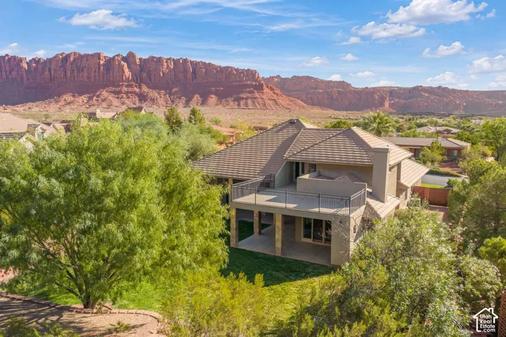 Multi-level residence featuring a tile roof, expansive upper-level metal-railing balcony, lower-level covered patio, and stone pillar accents, all set against a prominent red rock mountain backdrop