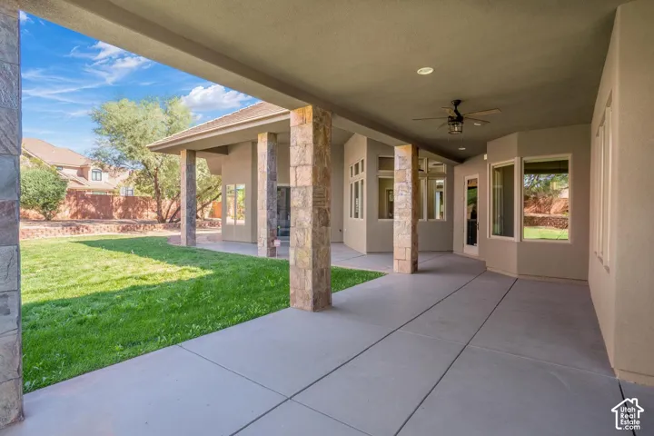 Covered patio featuring stone-clad support columns, a ceiling-mounted fan, concrete flooring, and windows overlooking the lawn