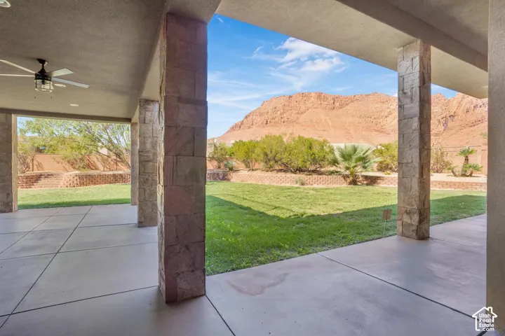 Covered patio featuring concrete flooring, stone-clad pillars, a ceiling fan, a grass lawn, and mountainous scenery