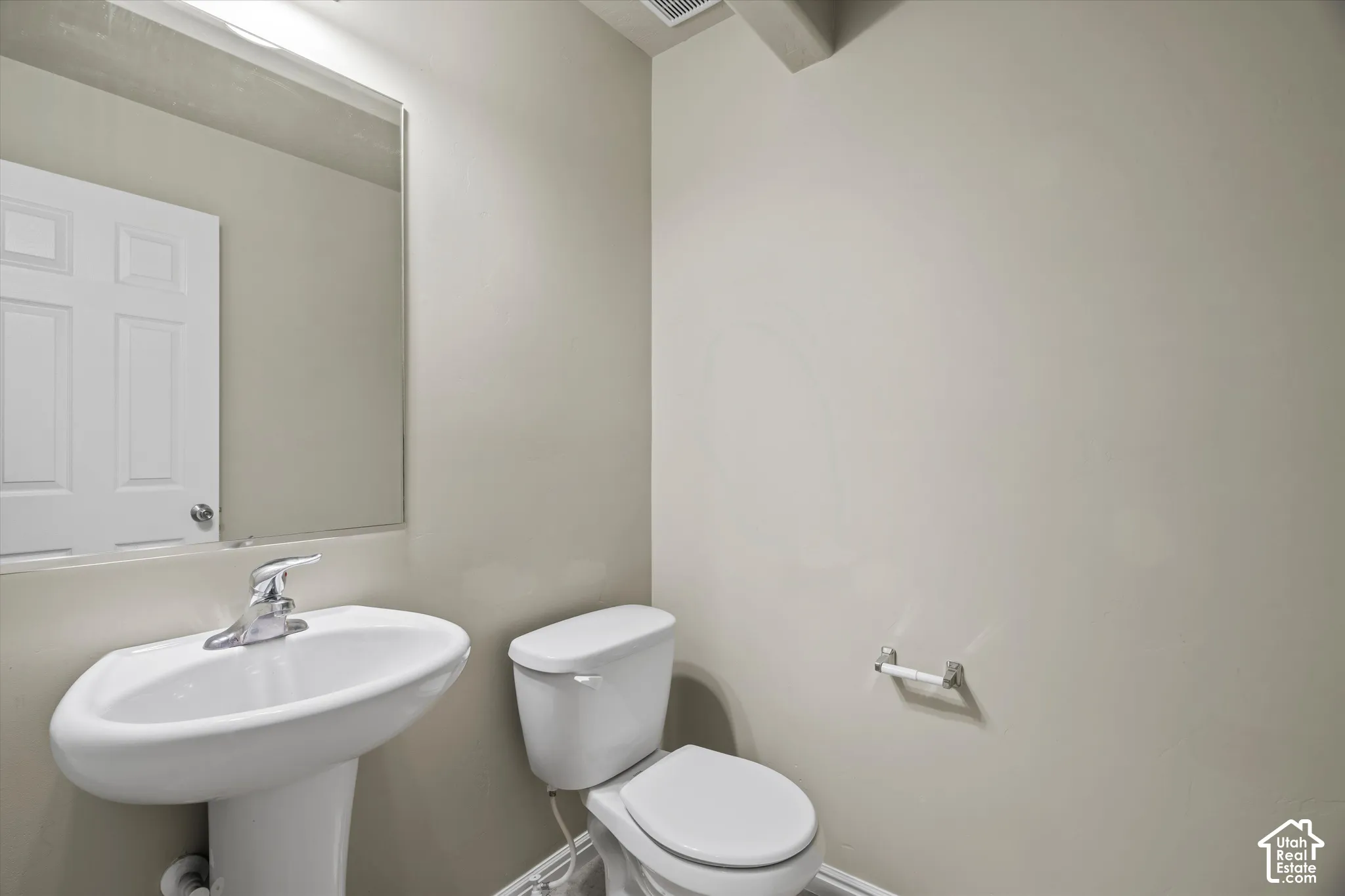 Wall-mounted frameless mirror above a white pedestal sink with chrome faucet and matching toilet