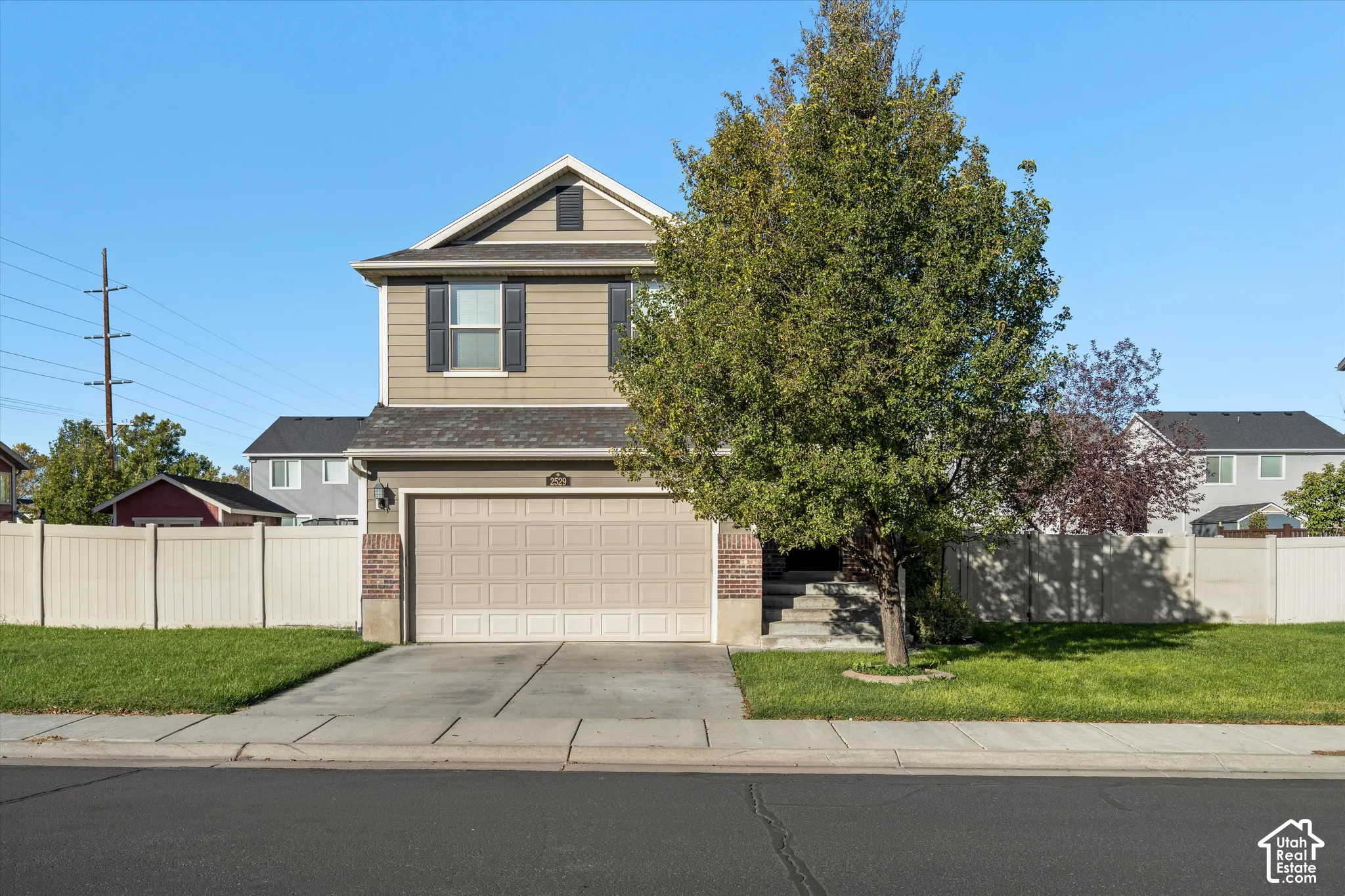 Two-story exterior featuring tan horizontal siding, a two-car garage, brick facade accents, and a fenced front yard with mature landscaping