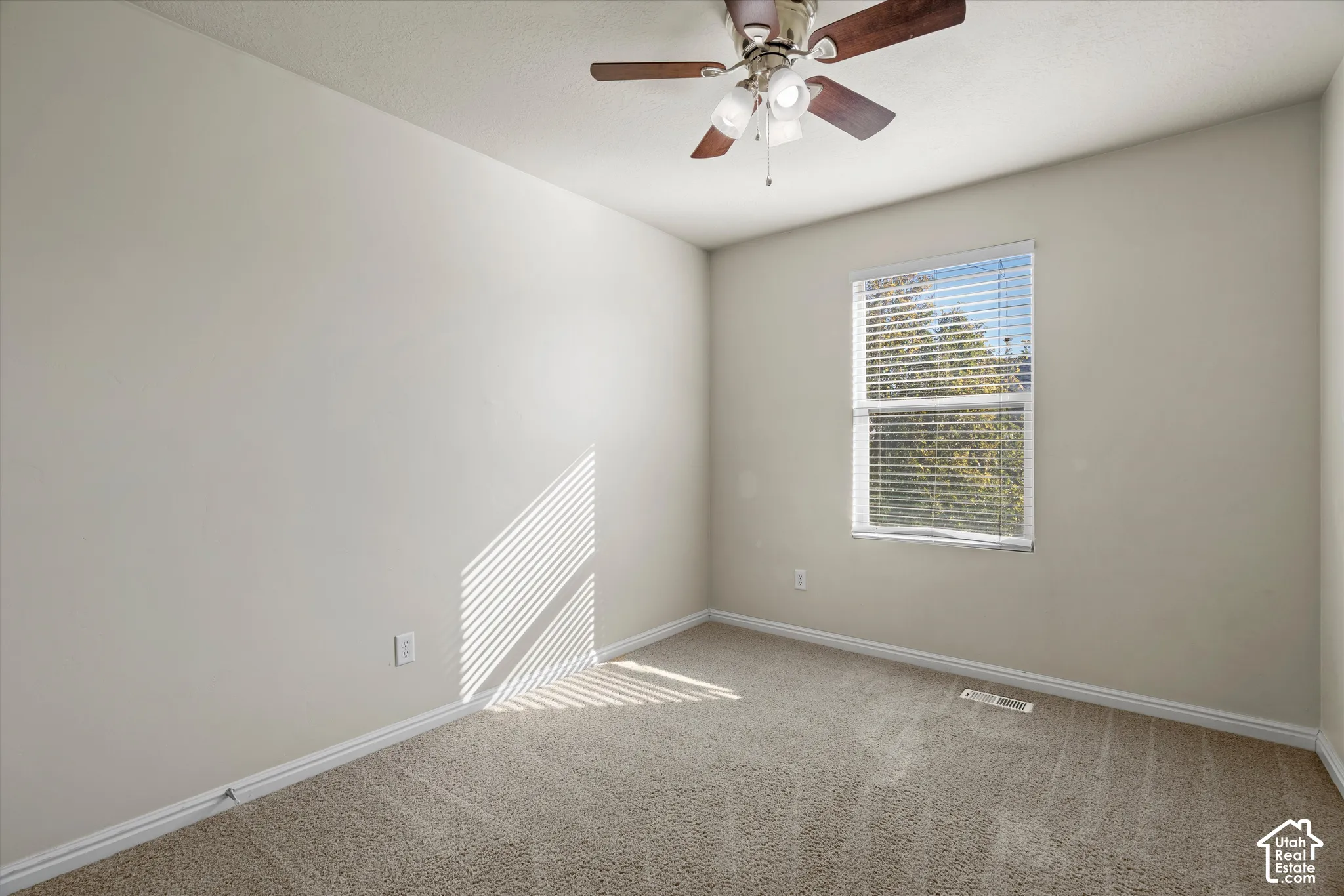 Neutral-toned interior space featuring textured wall-to-wall carpeting, a single-hung window with horizontal blinds, and a multi-blade ceiling fan with integrated lighting